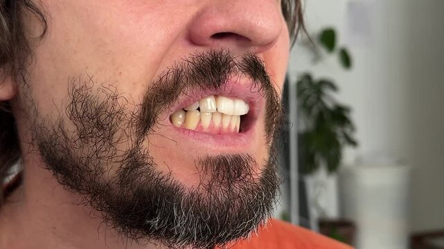 Close-up of man grinding his teeth with visible jaw tension. Beard and mustache enhance the intense facial gesture tied to stress, anger, or habit in indoor calm