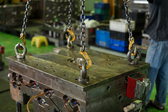 Multiple chain hooks secure heavy mold block from lifting eyes as worker prepares hoist movement, enabling safe transport, machine maintenance, and workflow control in manufacturing plant area