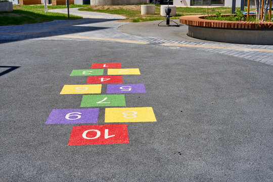 A colorful hopscotch game painted on an asphalt surface in an outdoor playground or park area. The hopscotch grid features ten numbered squares in alternating colors of red, green, yellow, and