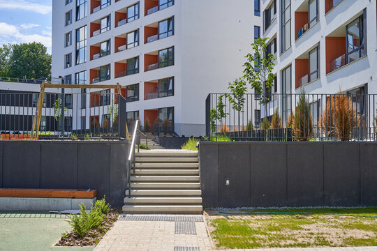 Modern residential apartment complex featuring multi-story buildings with white facades and orange-accented balconies. A central wide concrete staircase with metal handrails leads upward between