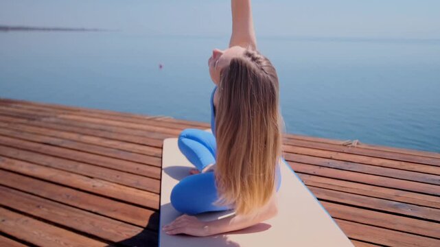 woman performing camel pose yoga side view leans back with arm reaching overhead on mat, wooden pier by lake, outdoor flexibility training, spine stretch control, strong posture during workout session