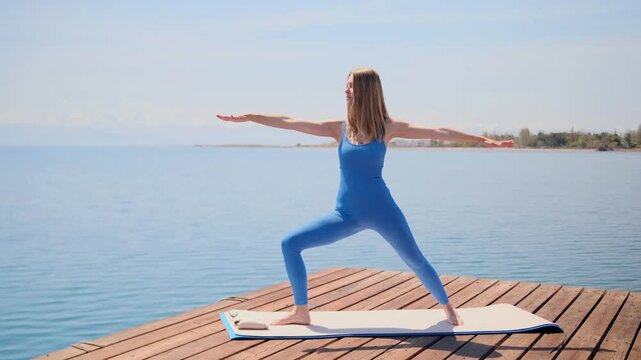 woman performing warrior ii yoga pose side view balances on mat on wooden pier by lake, outdoor fitness practice with strength stability and controlled posture during training session
