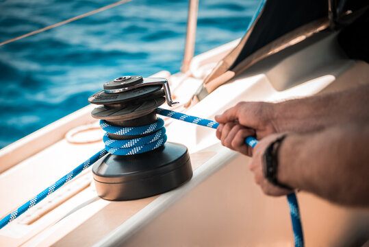 Sailing details, Man pulling a blue sheet/line on the winch with two hands, on a sunny day with blue water in the background, onbard a saildboat