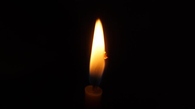 Bright candle flame close up on a deep black background. A single wax candle light flickering slowly in the dark