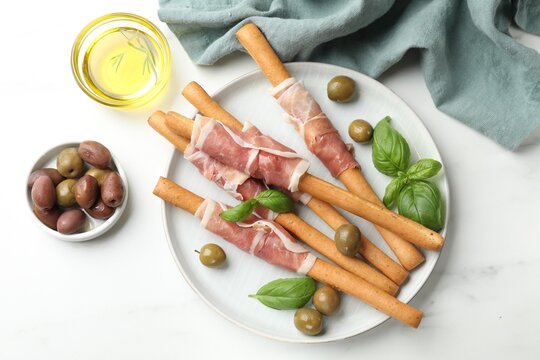 Grissini breadsticks with prosciutto, olives, oil and basil on white table, flat lay