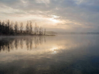Fototapeta premium Misty lake with tree reflections and soft sunrise light.