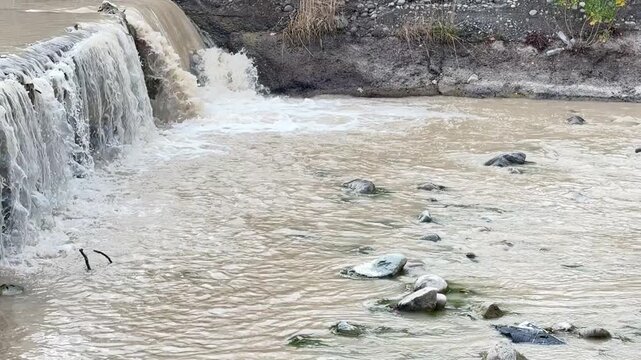 Water flowing over small dam into muddy river, urban drainage or stormwater runoff, strong stream movement, environmental concept, outdoor water flow scene