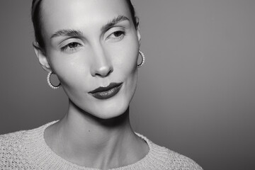 Fototapeta premium Close-up portrait of a slender Caucasian woman with light skin and short hair wearing earrings and a knitted sweater, looking sideways in a monochrome studio setting.