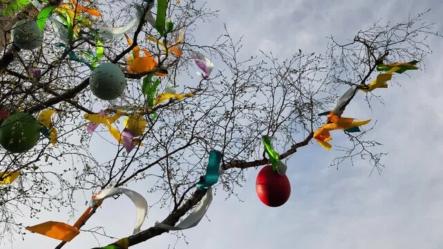 Bare tree branches decorated with colorful easter eggs and bright ribbons gently swaying in the wind against a cloudy spring sky
