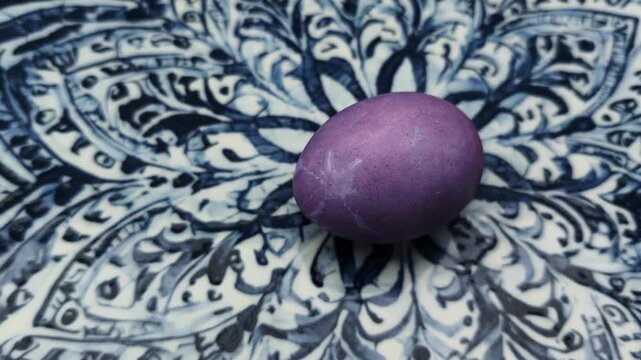 Single purple dyed easter egg rolling slowly across an ornate blue and white patterned plate. Traditional holiday symbol of spring and rebirth