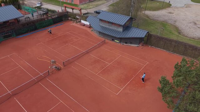 Two players engage in a tennis match on a clay court with a modern pavilion and surrounding greenery in the background