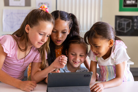 Diverse female children leaning around tablet folio case on classroom table studying cube poster