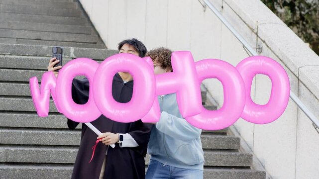 Two men on stairs, checking phone, lifting it, taking grad selfie, pink WOOHOO obscuring faces