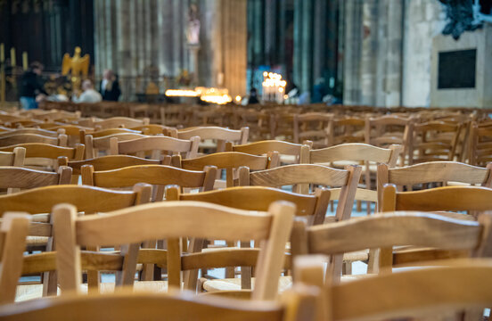 On a spring day in France, inside the Bordeaux Cathedral, wooden chairs for the parishioners are arranged in rows, creating a pattern. The chairs are made of light-colored wood with woven seats.