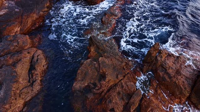 Top view of ocean waves breaking on coastal rocks. Vertical aerial shot of dark rocks and white sea foam in deep blue water.