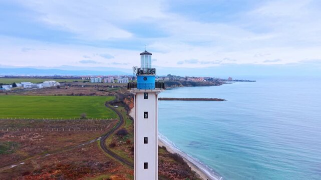 Close up of lighthouse tower with sea and fields. Detailed drone view of a white lighthouse beacon with green fields and blue sea in background.