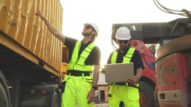Two engineers in safety vests analyze laptop data near cargo truck and container, coordinating logistics and industrial transport operations.