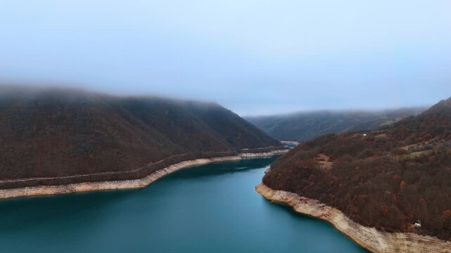 Turquoise Piva lake winding between steep fog covered hills. High angle view of a narrow river-like lake with white sediment shores and misty peaks in Montenegro.