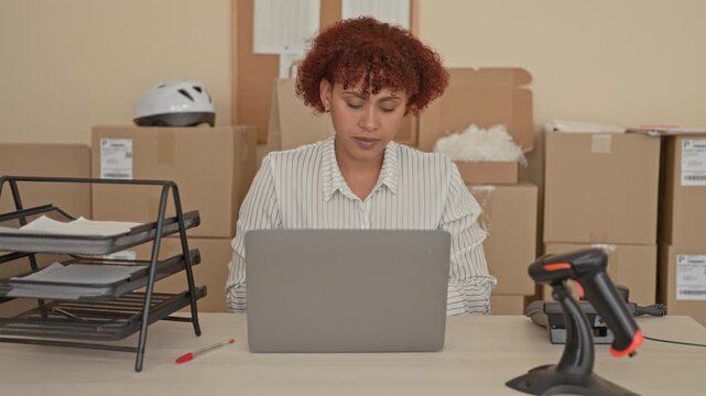 Woman closes laptop and stretches hands at a packing desk in a building surrounded by cardboard boxes, tray and barcode scanner; relief efficiency.