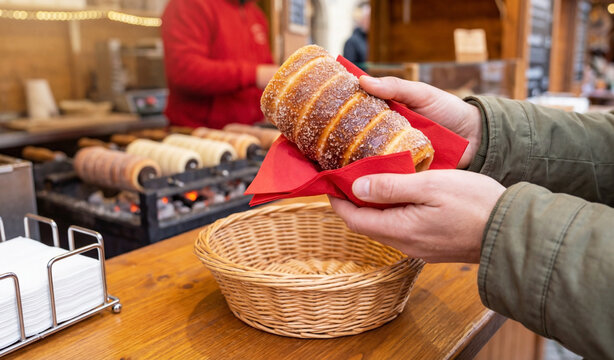 Close-up of hands holding a freshly baked Hungarian chimney cake, also known as k&uuml;rtőskal&aacute;cs or trdelnik, wrapped in a red napkin at a street food stall.