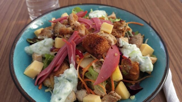 Close-up of a fresh mixed salad with grilled chicken pieces, pickled red onions, cheese cubes, croutons and creamy herb dressing served in a bowl on a wooden table. Bright, appetizing textures and col
