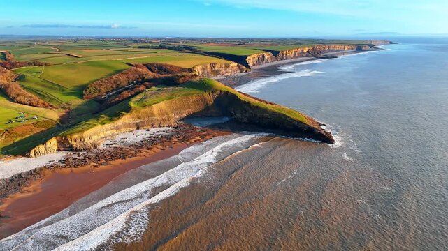 Wide aerial shot of coastal cliffs and shoreline landscape. Elevated view of limestone cliffs and agricultural land bordering the ocean with white waves on the beach.