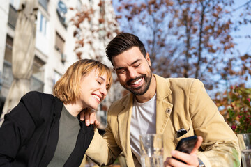 Fototapeta premium A happy young man and woman enjoy a sunny afternoon together at an outdoor city cafe, laughing while looking at content on a smartphone.