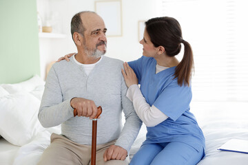 Fototapeta premium Care worker supporting senior man with walking cane on bed at nursing home