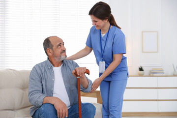 Obraz premium Smiling care worker supporting senior man with walking cane at nursing home