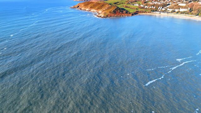 Aerial view of blue ocean waves reaching rocky Welsh coastline. High angle view of blue sea water approaching rugged cliffs and green hills in Wales during bright daylight.
