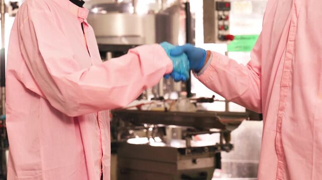 Two food production workers in protective clothing shaking hands inside clean manufacturing facility, symbolizing teamwork, hygiene standards, and cooperation