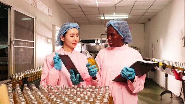 Food factory workers in protective clothing inspecting bottled beverages on production line, recording data for quality control and safety compliance