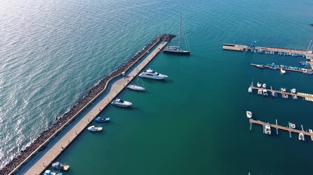 Long concrete pier and shorter berths with boats and yachts nearby. Drone flight over marina at daytime.