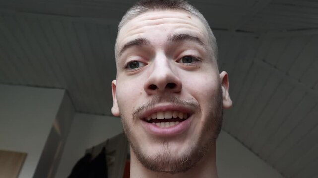 Low angle portrait of young Gen Z man looking down at camera, talking on video call or reacting to smartphone content, casual vlog