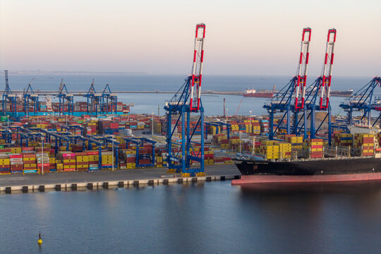 Gdansk, Poland - 27 March 2026: Aerial view of the container terminal at the Port of Gdansk with large gantry cranes, stacked shipping containers, and a cargo ship docked at the pier during sunset.