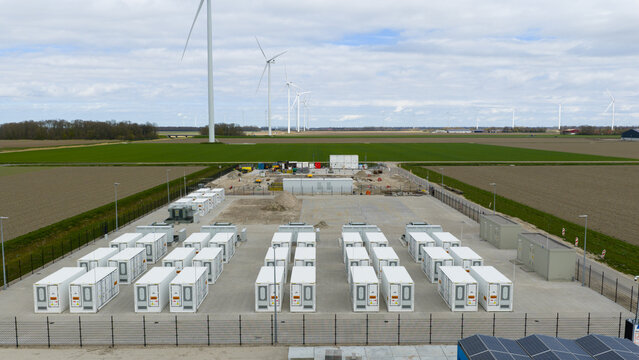 Dronten, Netherlands - 04 April 2026: Aerial view of the Olsterdwarspad battery energy storage system facility with rows of white containers and wind turbines in the background.