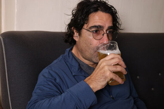 man relaxing at home sitting in a chair reading a book and drinking a lager beer from glass (couch living room)