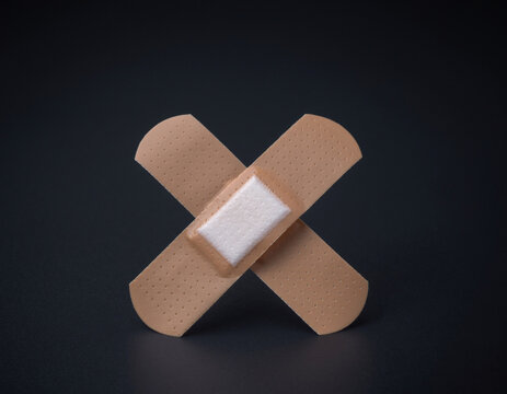 Two adhesive bandages are placed in an X formation on a dark background. These medical plasters are for wound care and are part of first aid supplies for cuts and scrapes. They symbolize the need for 