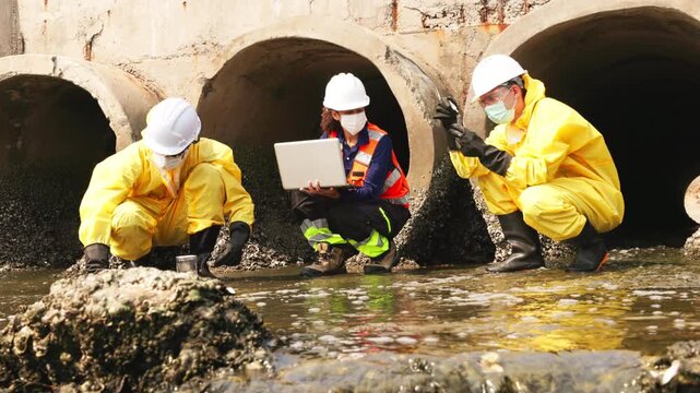 Group of environmental workers in protective suits collect and analyze water samples near drainage pipes, monitoring chemical contamination and industrial wastewater pollution.