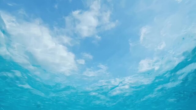 Underwater view looking upward through transparent turquoise water toward sunlit sky and clouds, showing rippling surface and light refraction.