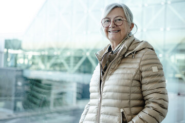 Smiling senior woman stands by a large airport terminal window wearing a beige puffer jacket and glasses, conveying confidence, travel readiness and stylish comfort in a bright modern setting. © luciano