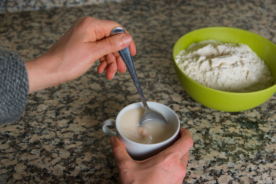 Manos de mujer revuelven con una cuchara de metal levadura de panader&iacute;a en una taza con agua, con un cuenco con harina dentro de fondo