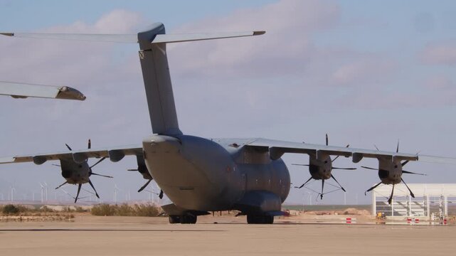 ZARAGOZA, SPAIN - FEBRUARY 23, 2026: Rear view of a large Airbus A400M military transport aircraft parked on a hot airfield tarmac. Wind turbines are visible on the horizon under a partly cloudy sky