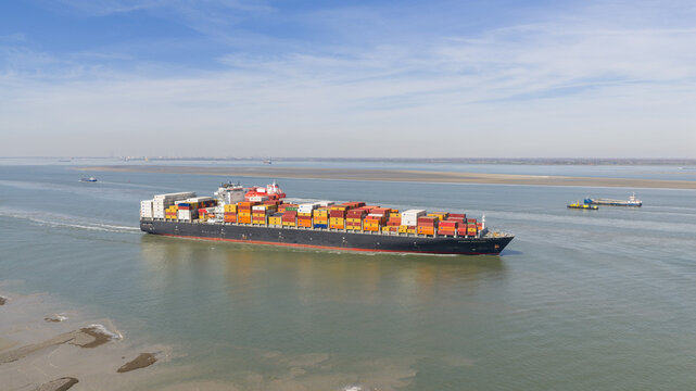 Terneuzen, Netherlands - 08 April 2026: Aerial view of a large container ship navigating the Westerschelde estuary with colorful cargo containers under a clear blue sky.