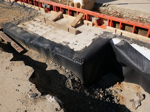  A high-angle view shows a black HDPE dimpled drainage
membrane being installed along a concrete foundation wall at a construction
site. Red modular formwork panels and wooden supports are visible in 