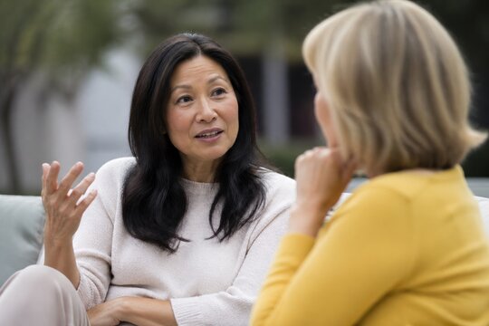 Two mature women sit on a sofa outdoors talking. They share a serious and engaged moment with natural lighting and a relaxed atmosphere. Perfect for mental health or friendship articles.