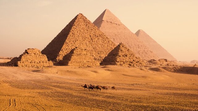 Group camels and their owners on desert plateau with Pyramids of Giza in soft morning light, Egypt. A group camels with their owners walks across sandy desert plateau near Pyramids of Giza at sunrise