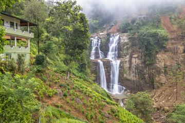 Ramboda Waterfall near Nuwara Eliya town in Sri Lanka. Majestic multi-tiered waterfall cascades through lush green mountainside with a charming hillside dwelling nearby © Viliam