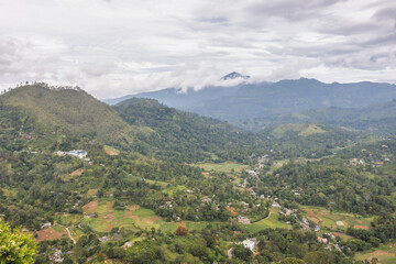 Fototapeta premium Panoramic view of lush green mountain landscape with distant peaks shrouded in clouds under a dramatic sky. Mountain landscape near Ella town in Sri Lanka island in Asia.