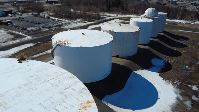 Aerial drone footage of dome‑topped oil storage tanks, pipelines, and energy infrastructure casting long shadows across snowy industrial grounds in Matane, Quebec, Canada.